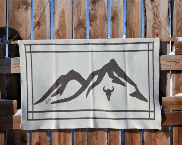 The 1889 Limited Edition Montana blanket hanging from a wooden fence showing the woven Montana mountains and cow skull.