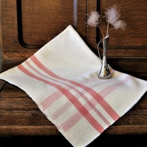Baby blanket with a light red cross-weave pattern folded and resting on a piece of furniture with a small silver vase and three flowers placed on top.