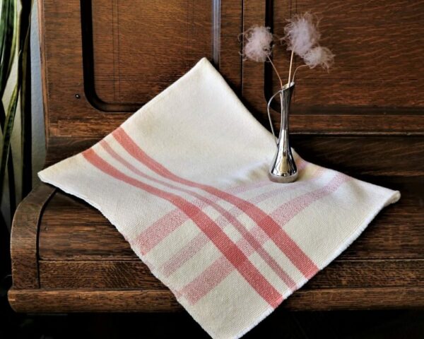 Baby blanket with a light red cross-weave pattern folded and resting on a piece of furniture with a small silver vase and three flowers placed on top.